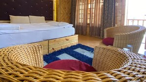 Interior de una cabaña de madera con una cama, sillas de mimbre y un ventanal con cortinas.