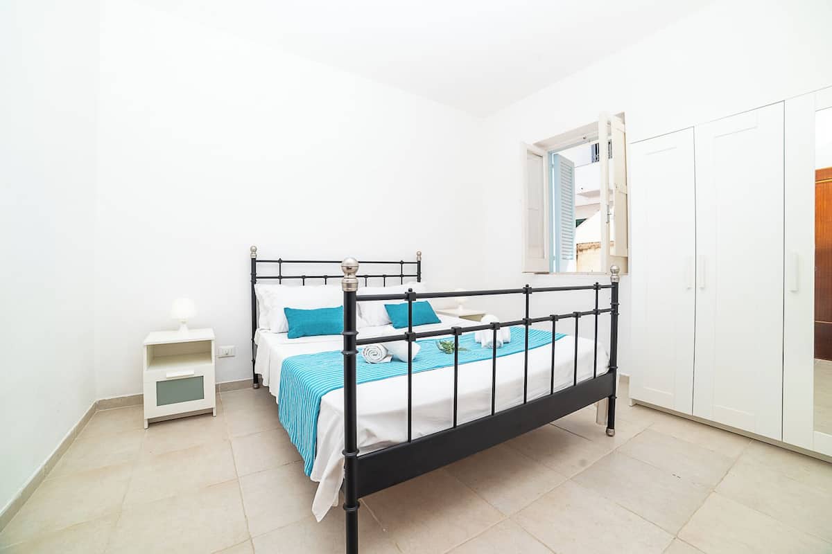 A single bed with a black metal frame, white bedding, and blue accents. There is a small bedside table with a lamp, and a white wardrobe with a mirror.