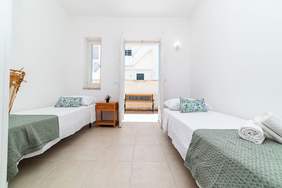A small, bright room with two single beds, a small wooden nightstand, and a window.
