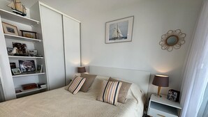 A neatly arranged bedroom with a large bed, a bedside table, a nightstand, a bookshelf, and a framed picture on the wall. - Charming 2-room apartment with pool in Hendaye, near port (Hendaye)