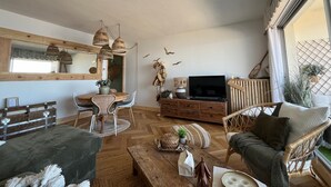 A living room with a wooden floor, wicker furniture, a television, and a large window. - Arcachon - 2-Bedroom Apartment with Terrace, Sea View and Parking (Arcachon)