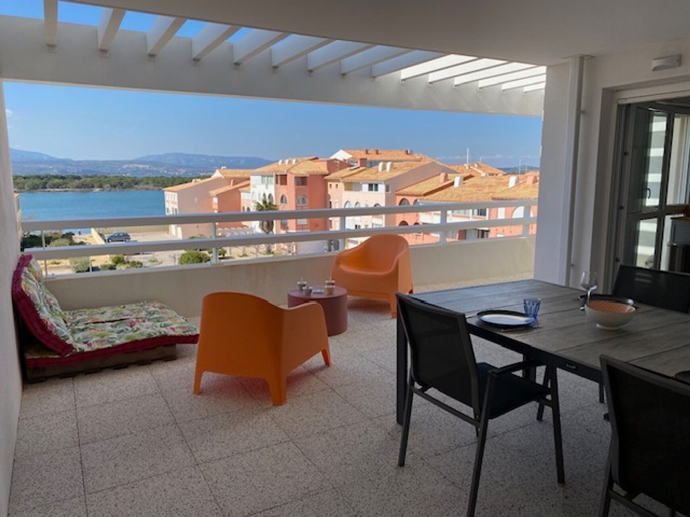 A balcony with a table, chairs, and a sofa, overlooking a coastal area with buildings and a clear sky.