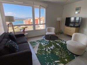 A living room with a sofa, armchair, coffee table, and a TV. There is a large window with a view of buildings and a body of water.