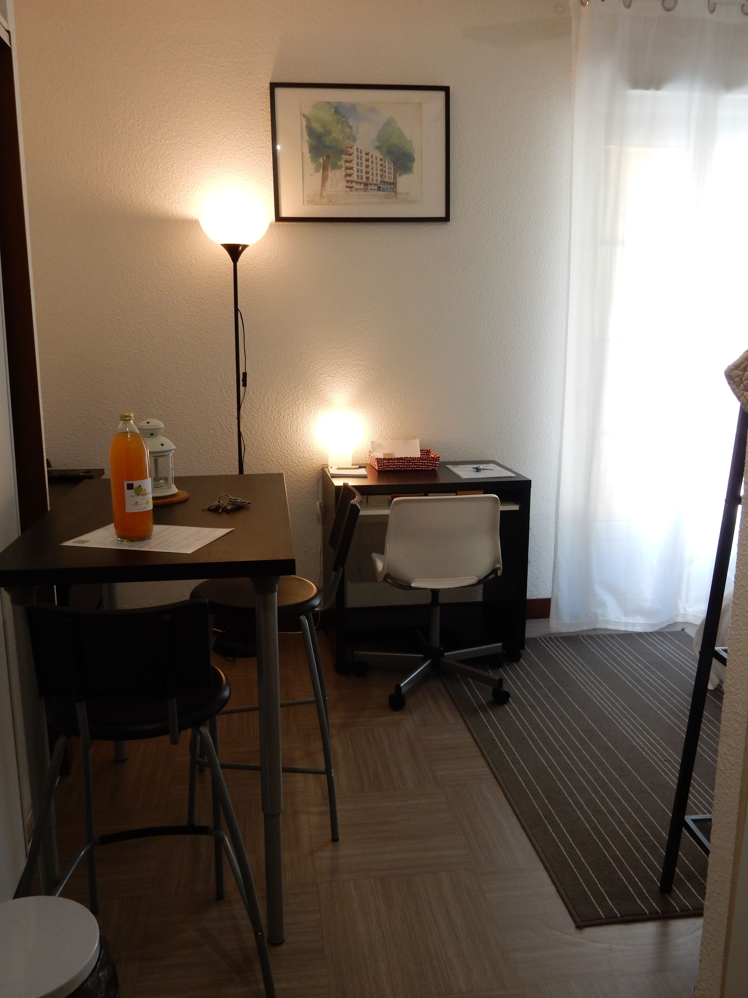 Une chambre d’hôtel avec un bureau en bois, une chaise, une lampe, un tableau encadré au mur et une fenêtre avec un rideau.