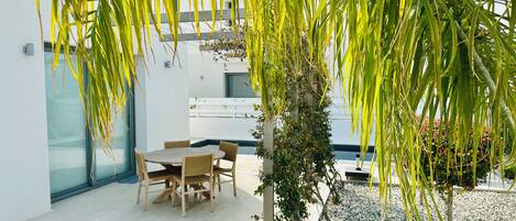 A modern outdoor dining area with a table and chairs, surrounded by greenery and a white wall.