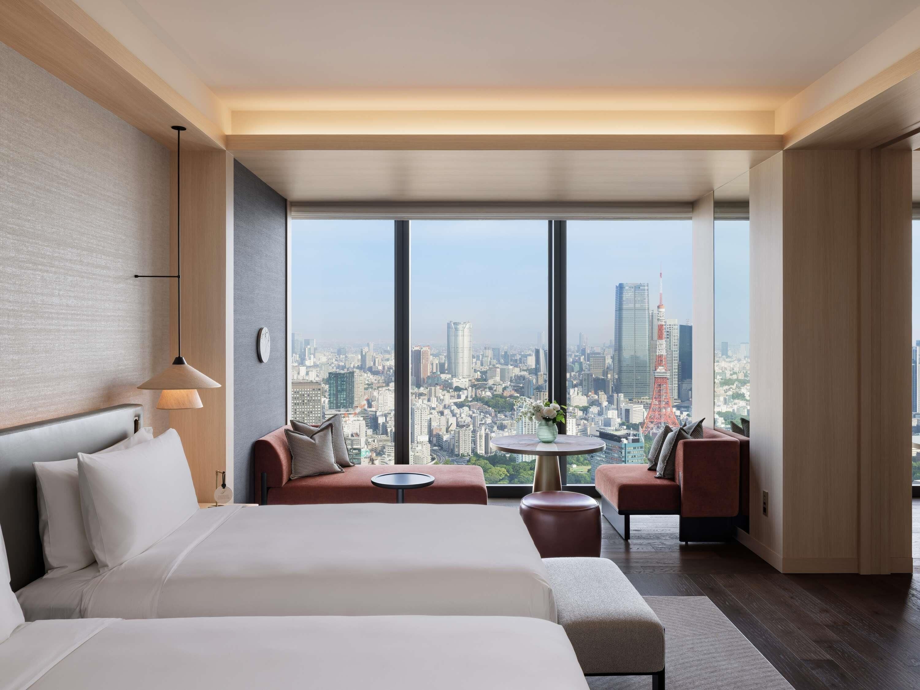 A modern hotel room with a large bed, a seating area with a table, and a city view through large windows.