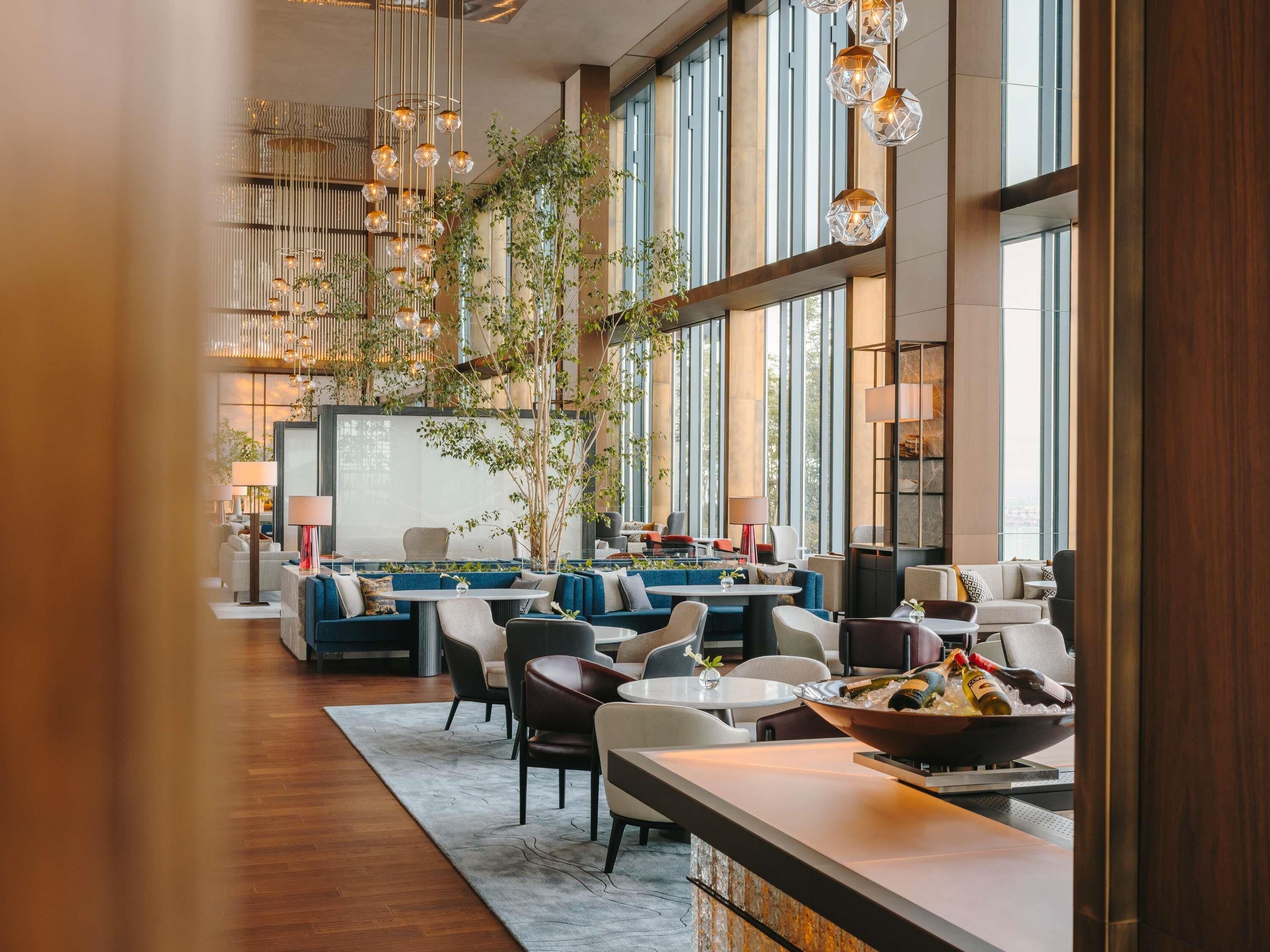 A modern hotel lobby with a central seating area, large windows, and hanging light fixtures.