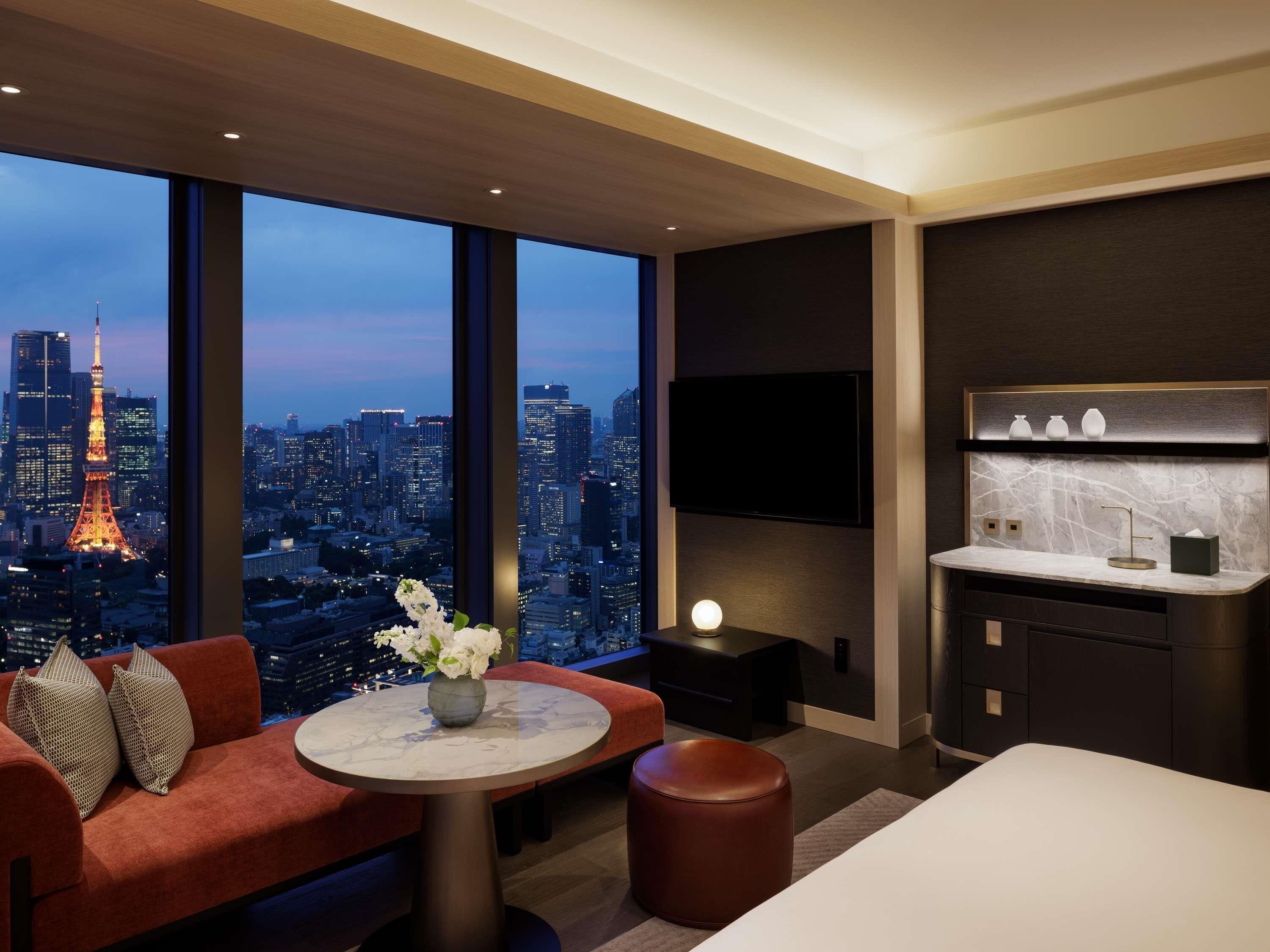 A modern hotel room with a large window offering a city view, a flat-screen TV mounted on the wall, a round marble table, and a cozy seating area with a sofa and ottoman.