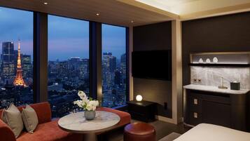 A modern hotel room with a large window offering a city view, a flat-screen TV mounted on the wall, a round marble table, and a cozy seating area with a sofa and ottoman.