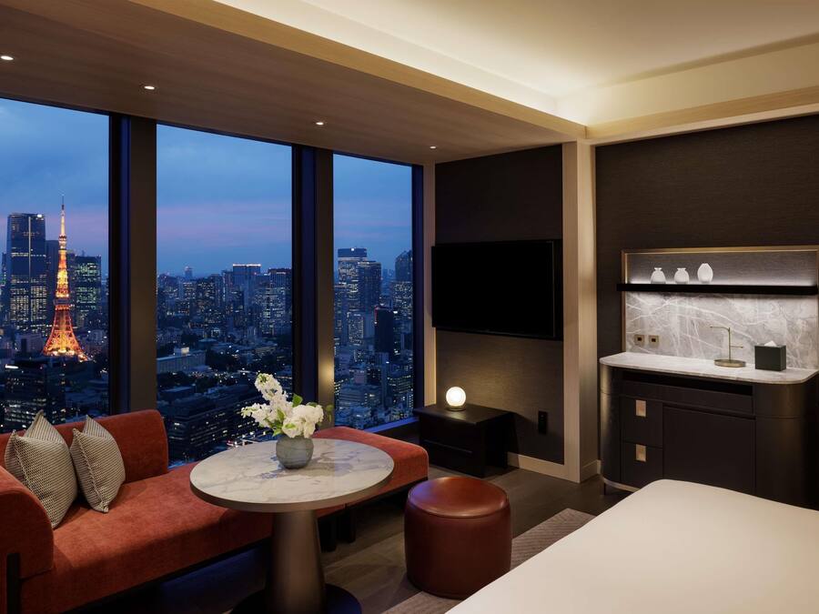 A modern hotel room with a large window offering a city view, a flat-screen TV mounted on the wall, a round marble table, and a cozy seating area with a sofa and ottoman.