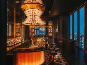 Uma área de bar com teto alto, um lustre grande e vista para a cidade através de amplas janelas.