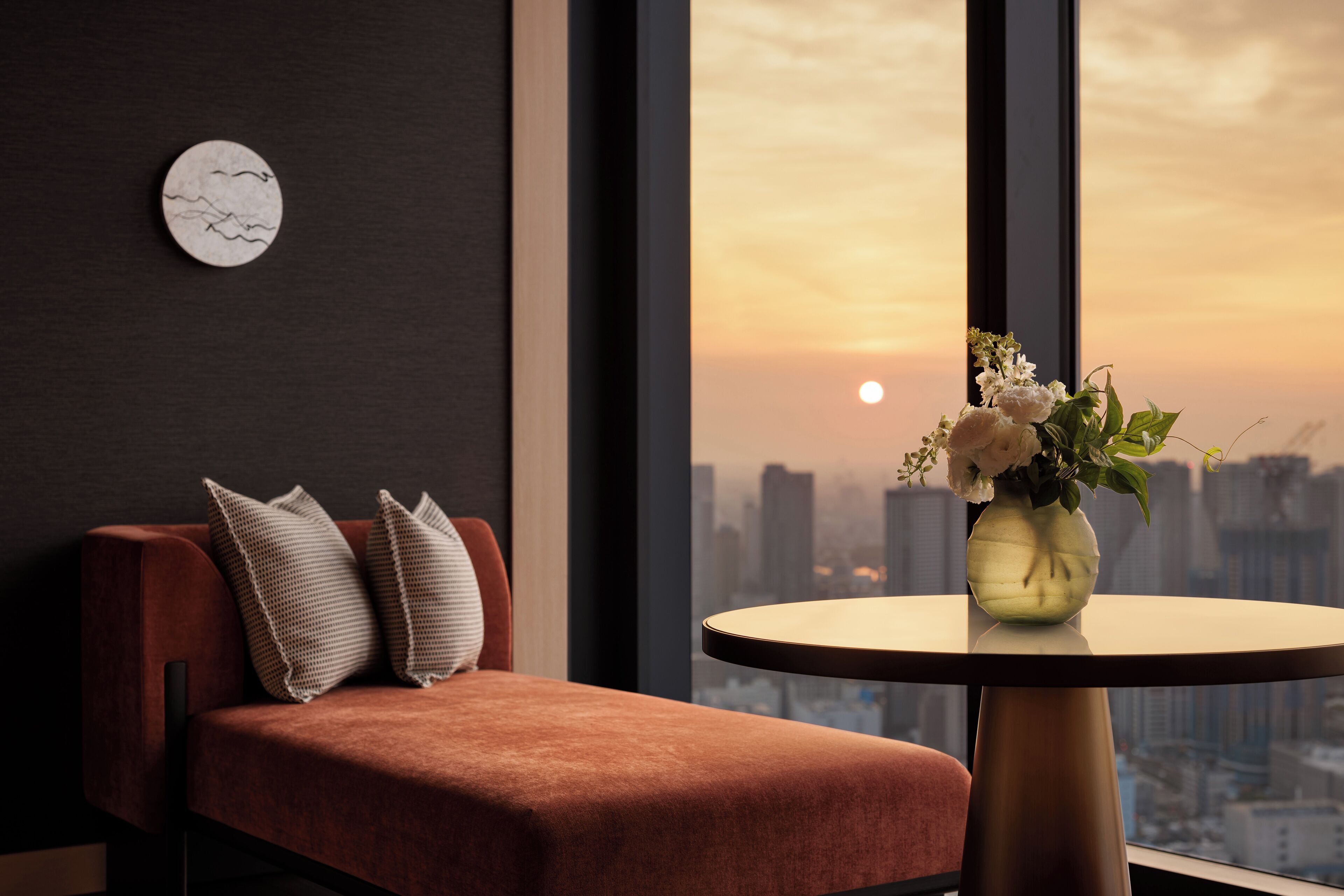 A modern room with a red sofa, a round table with a vase of flowers, and a cityscape view at sunset.