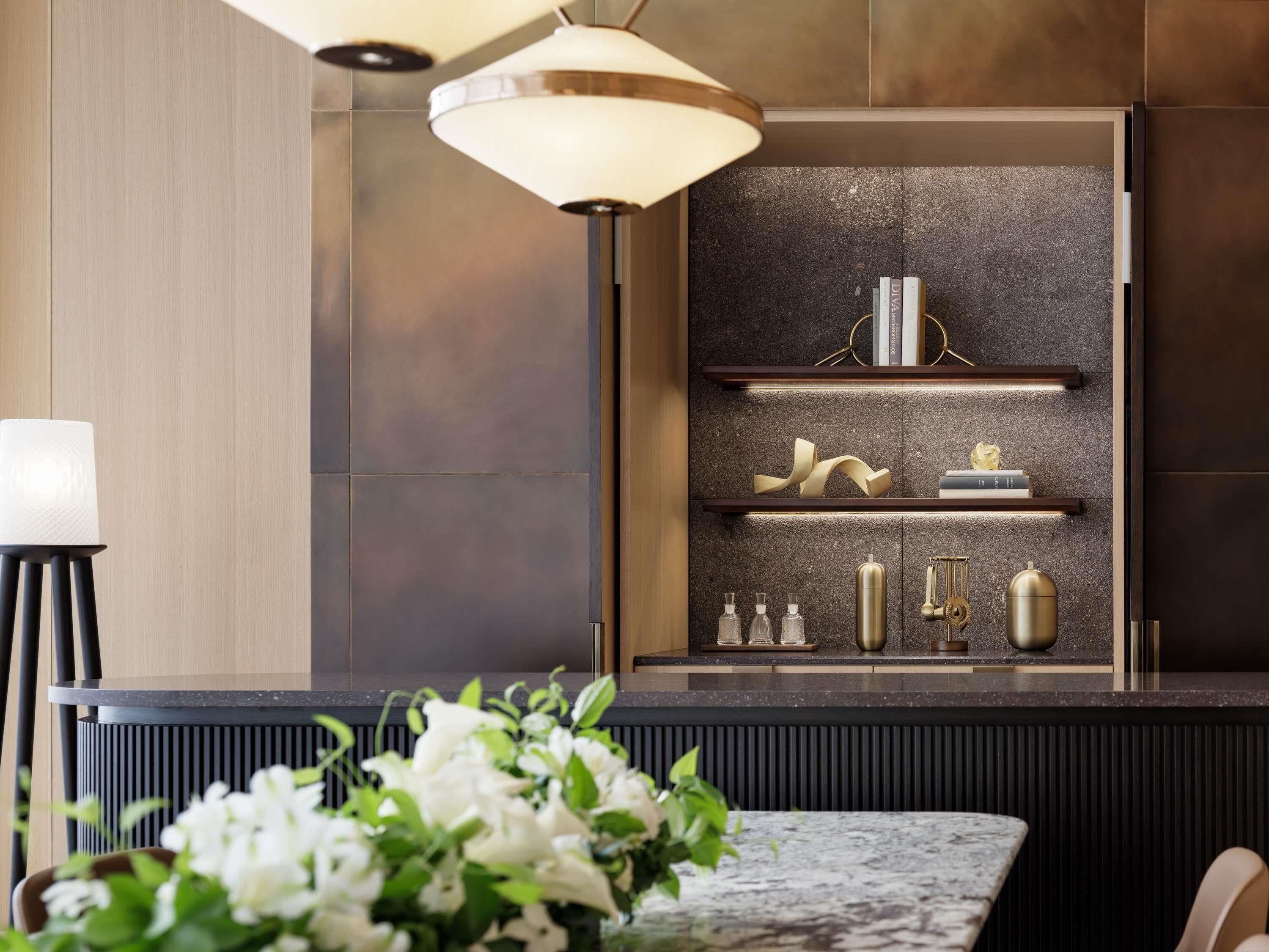 Una cocina moderna con salpicadero oscuro, armarios de madera y encimera de mármol. Se observan estantes con objetos decorativos y un jarrón alto y cilíndrico.