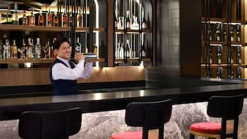 A bar area with a bartender, shelves of liquor bottles, and bar stools with red cushions.