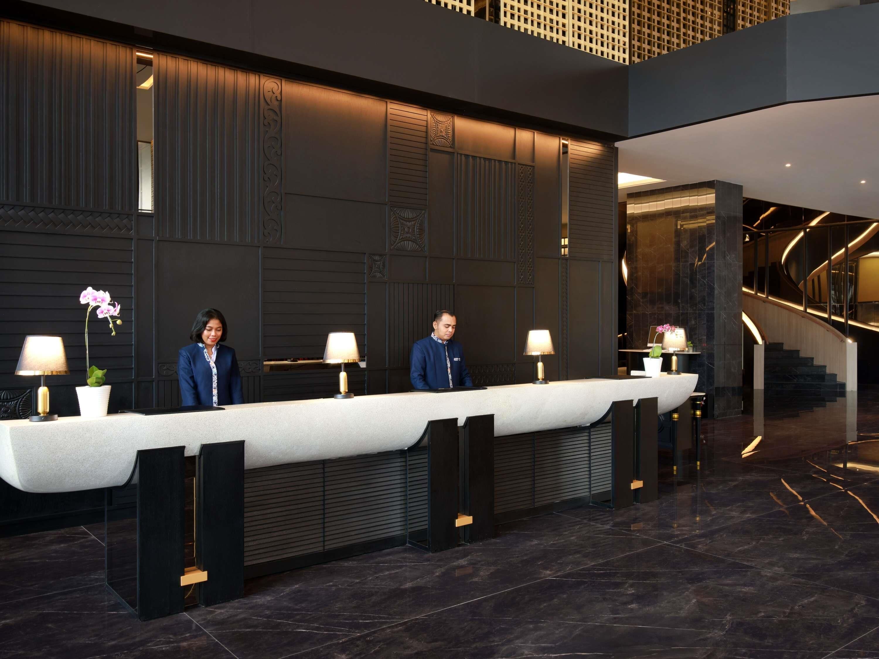A reception desk with two staff members, a modern interior with dark walls and a marble floor, and a staircase in the background.