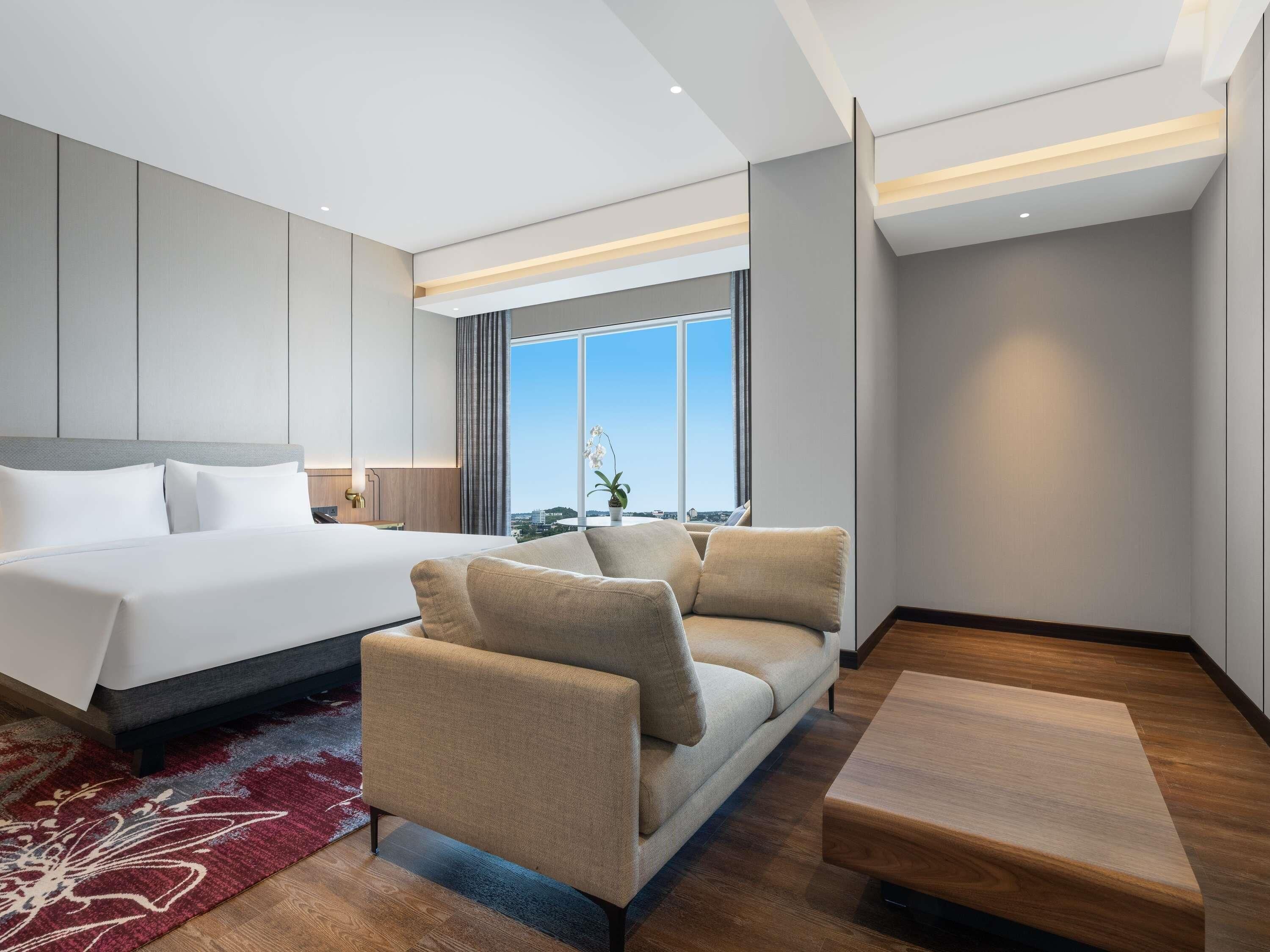 A modern hotel room with a large bed, a sofa, a small table, and a view of the city.