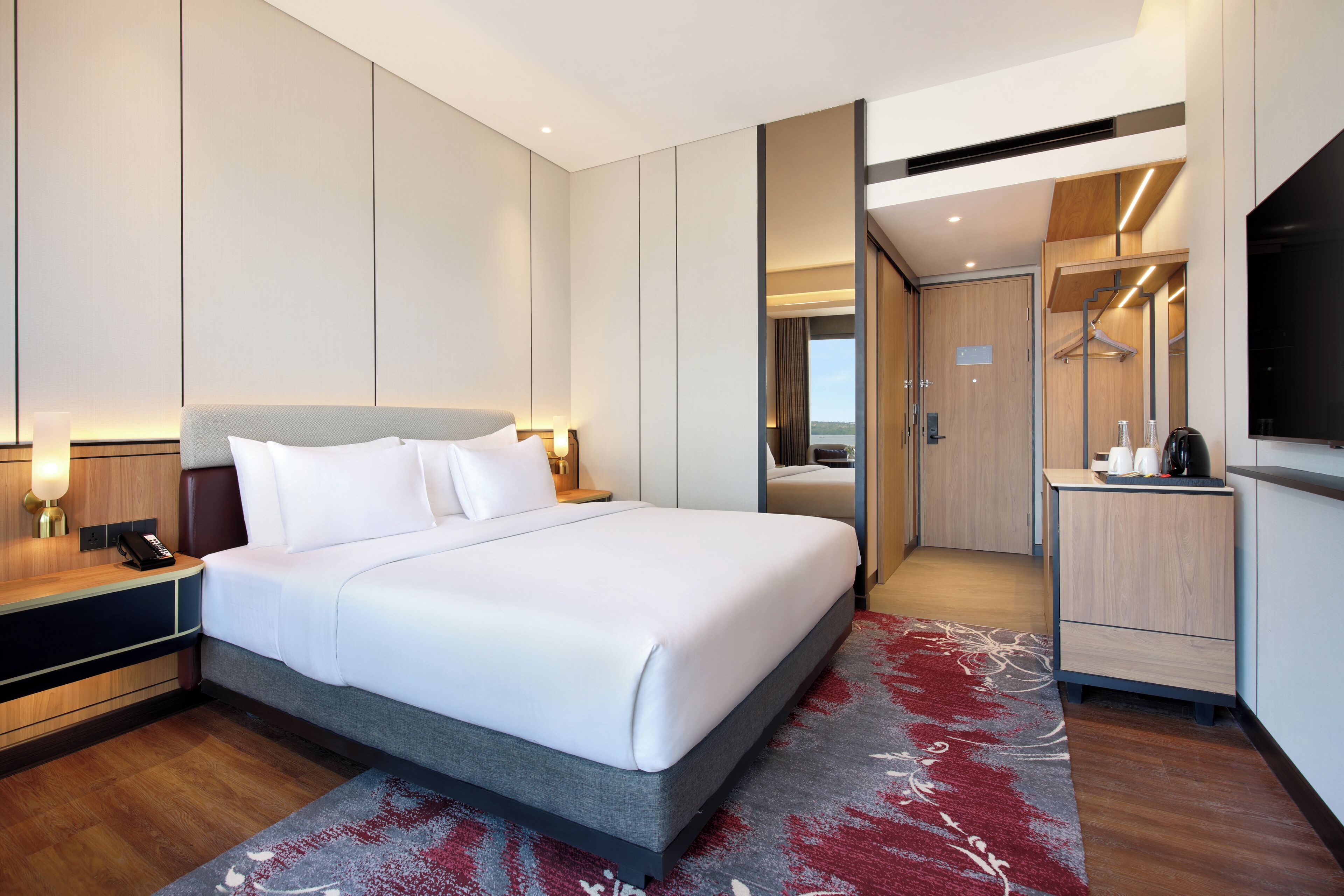 Et moderne hotelværelse med en stor seng, et natbord med lampe, et spejl, et klædeskab og et fjernsyn.