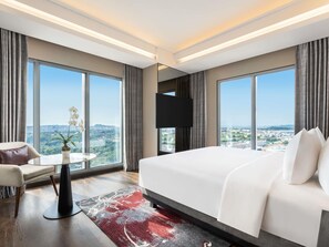 A hotel room with a large bed, a TV, a small table with a chair, and a view of a cityscape through large windows. - Grand Mercure Batam Centre (Batam)
