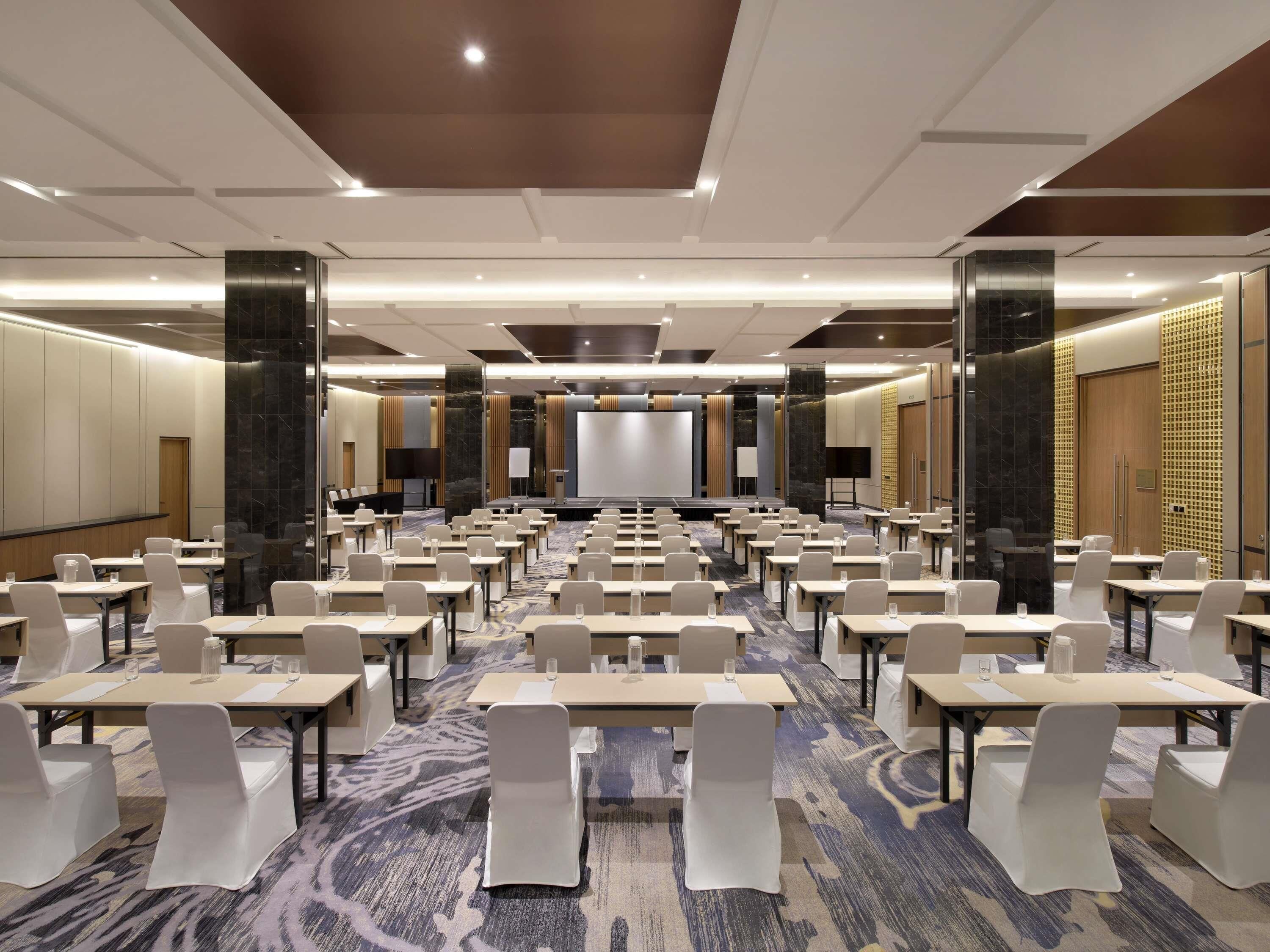 A conference room with rows of tables and chairs, a stage, and a large screen.