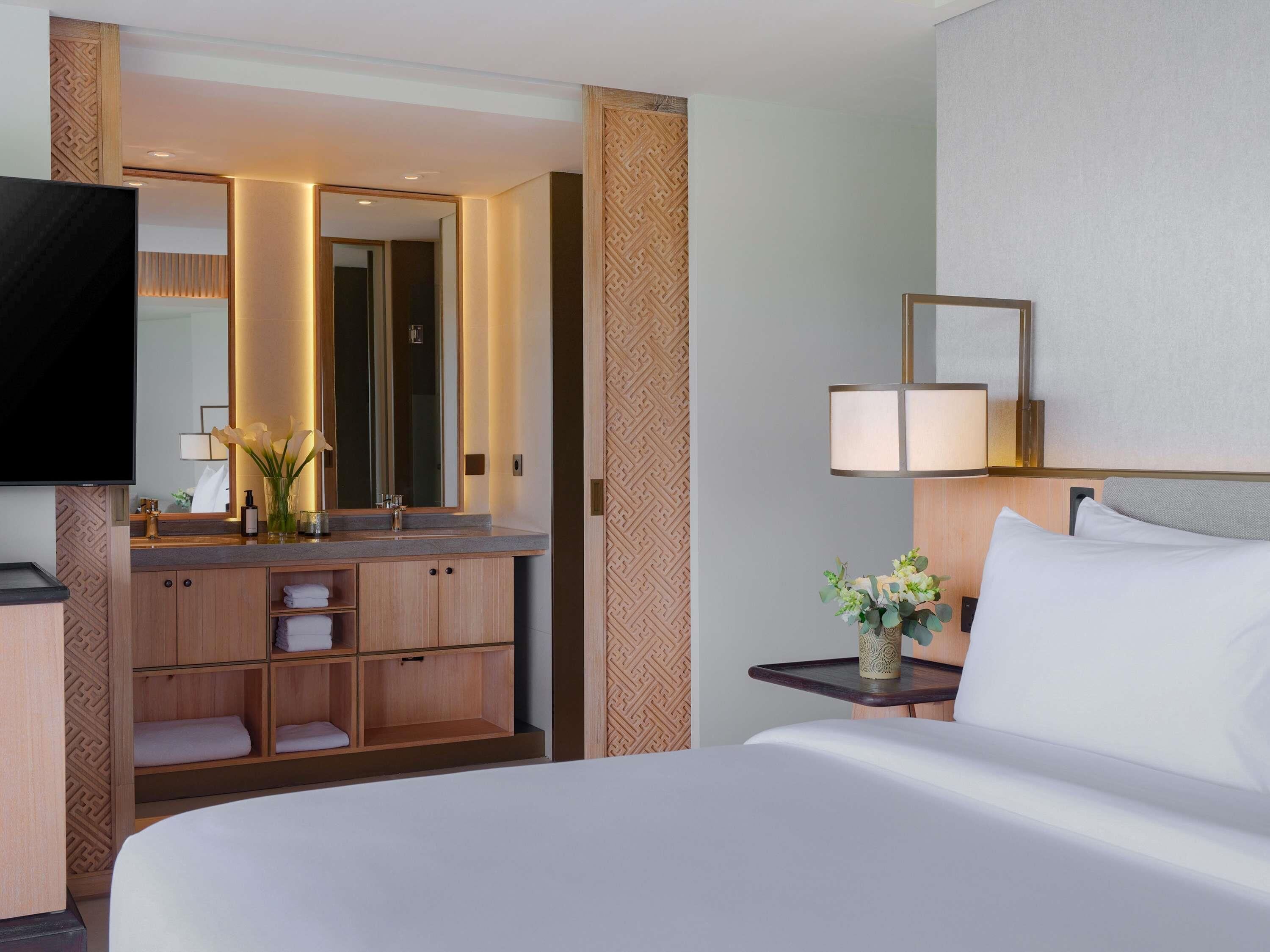 Une chambre d’hôtel avec un grand lit, une table de chevet avec une lampe, une télévision, une coiffeuse en bois avec un miroir, et une salle de bain visible à travers une porte ouverte.