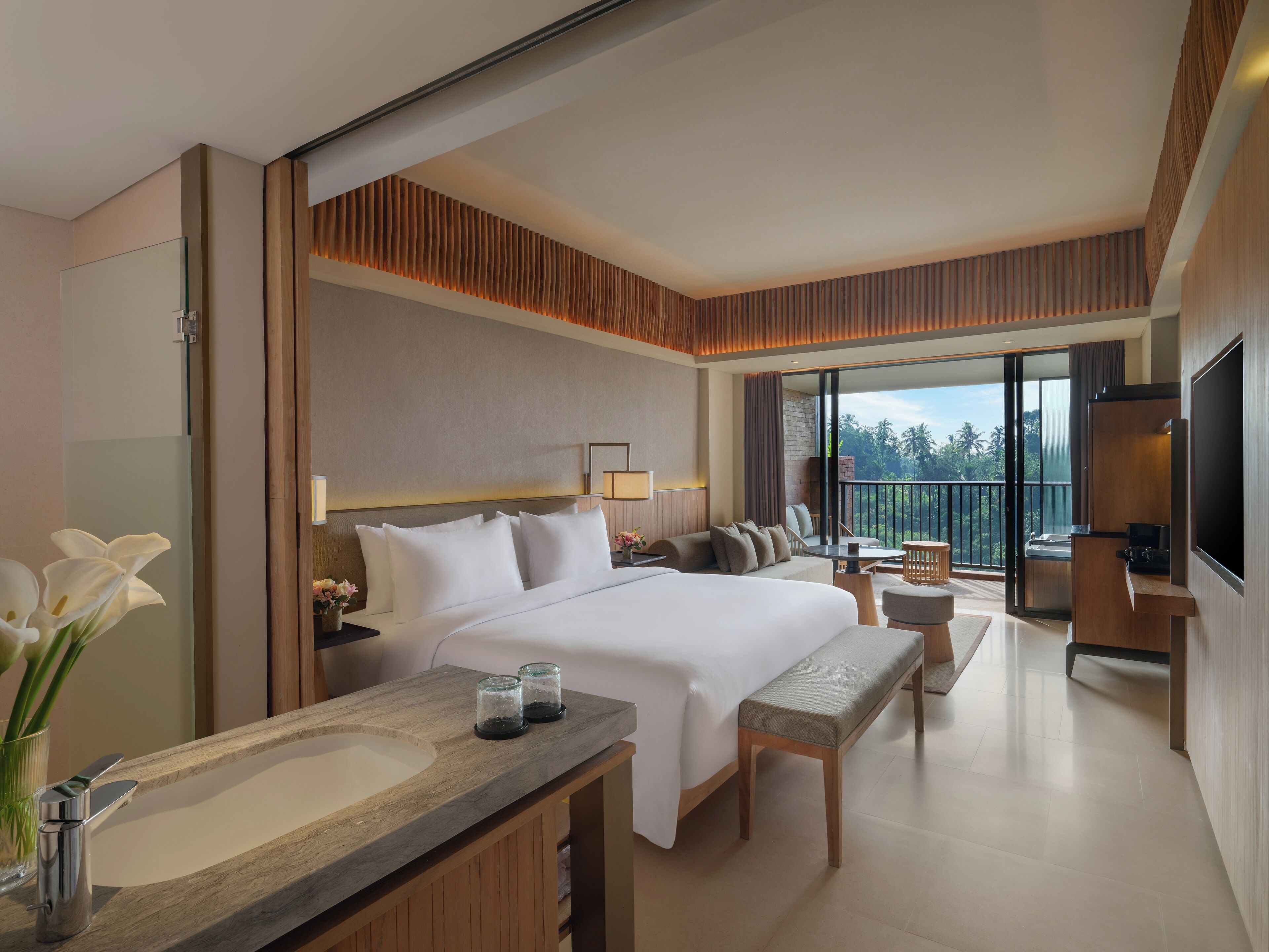 A modern hotel room with a large bed, a sitting area, a balcony with a view, and a TV.