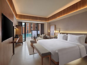 A modern hotel room with a large bed, a flat-screen TV, a desk, and a view of a swimming pool and building. - Novotel Bali Ubud Resort (Payangan)