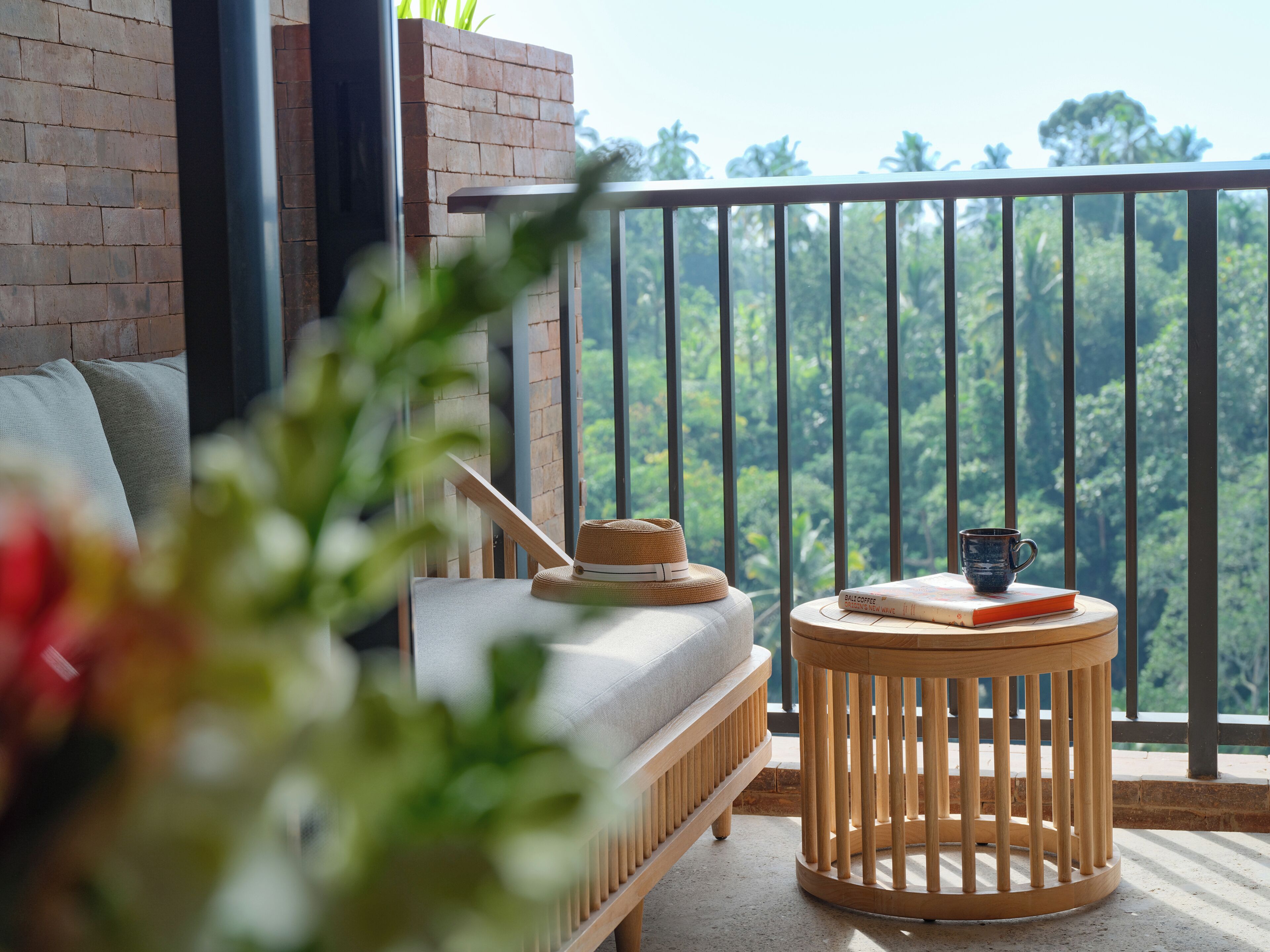 A balcony with a view of a lush forest, a round wooden table with a book and a cup, and a sofa with a hat on it.