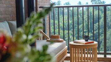 A balcony with a view of a lush forest, a round wooden table with a book and a cup, and a sofa with a hat on it.