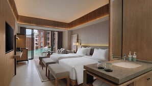 A hotel room with two beds, a large mirror, a desk with a chair, a small table, and a view of a pool and buildings outside. - Novotel Bali Ubud Resort (Payangan)