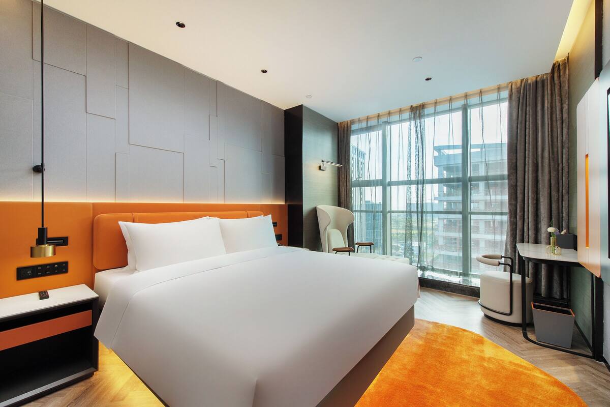 A modern hotel room with a large bed, a nightstand, a desk, a chair, and a large window with curtains.