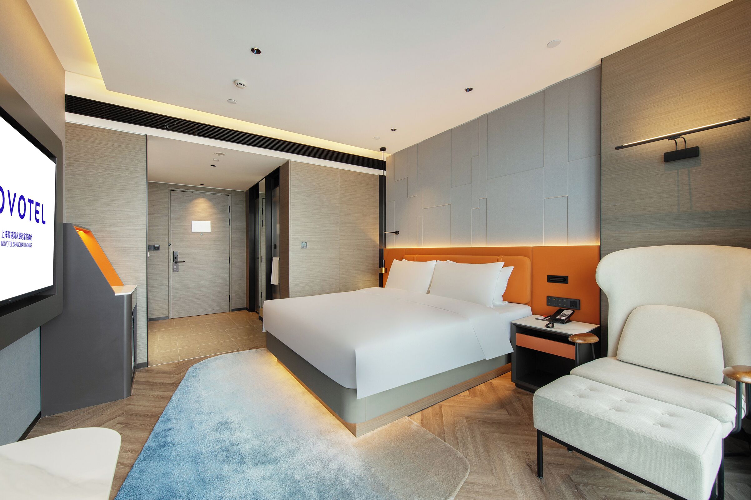 Ein modernes Hotelzimmer mit einem großen Bett, einer Sitzecke mit einem Sessel, einem Schreibtisch und einem Fernseher, der Hotelinformationen anzeigt.