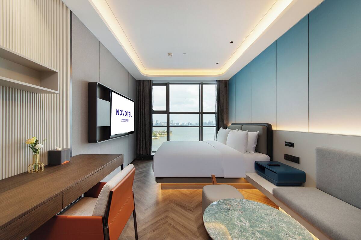 A modern hotel room with a large bed, a desk with a chair, a sofa, and a TV mounted on the wall.