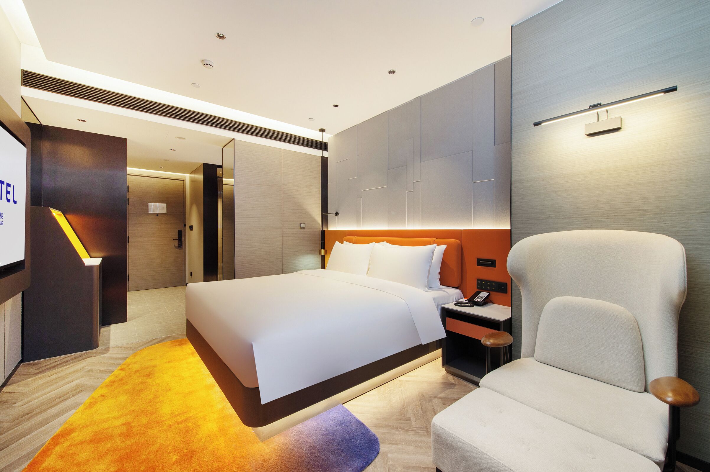 A modern hotel room with a large bed, a seating area with a chair, a desk with a lamp, and a wall-mounted light fixture.