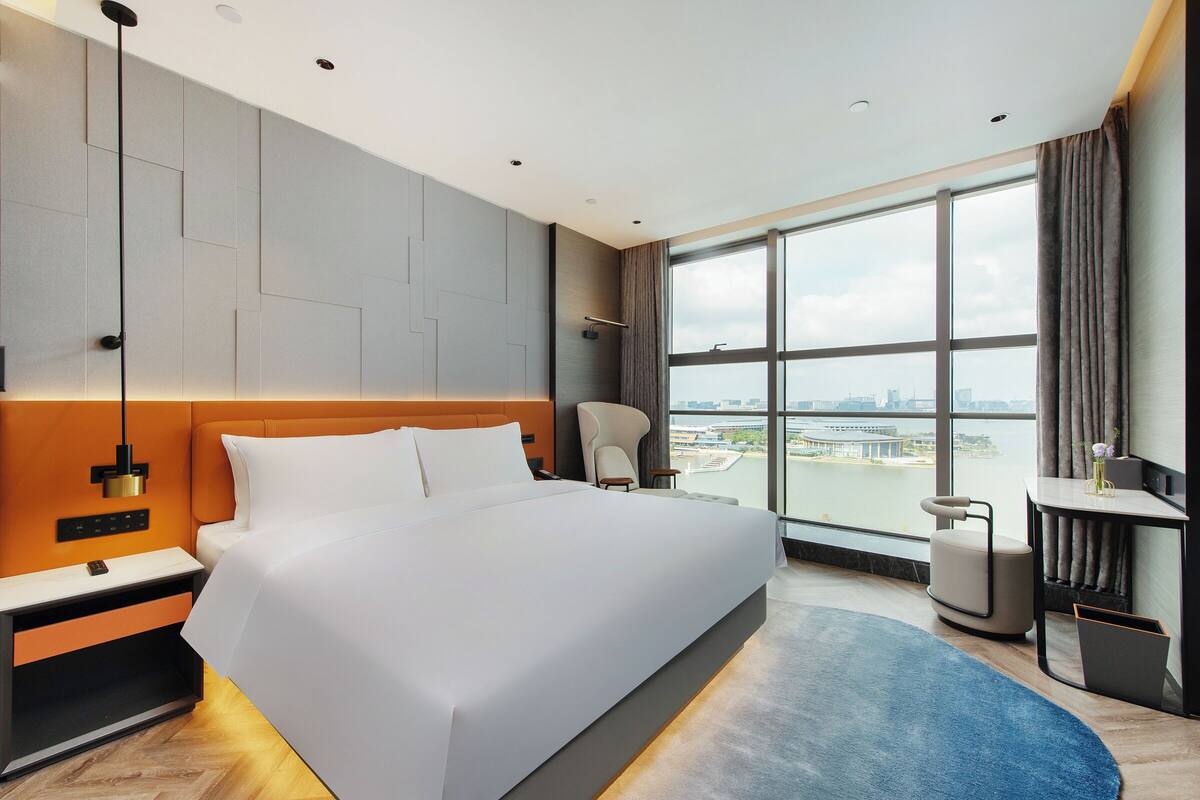 A modern hotel room with a large bed, a nightstand, a desk, and a view of the city through large windows.