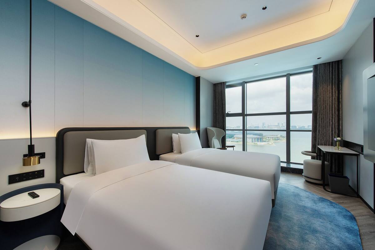A modern hotel room with two beds, a desk, a chair, and a large window offering a city view.