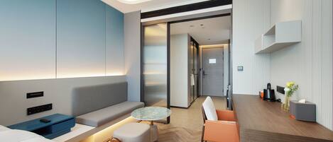 A modern hotel room with a sofa, a small round table, a coffee table, a dining area with a table and chairs, and a kitchenette.