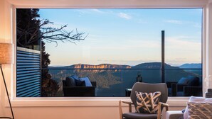 Una habitación con un ventanal grande que ofrece vistas a un lago y montañas. Hay una silla con cojín, una mesita redonda pequeña y un sofá con cojines.