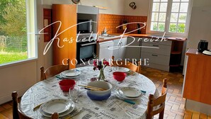 A dining area with a round table set for four, surrounded by chairs. A kitchen with a window and a door is visible in the background. - Le Gîte des Sabotiers by Kastell Breizh (Canihuel)