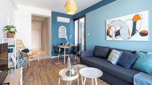 A modern living room with a blue accent wall, a gray sofa, a dining area with a round table, and a large abstract painting. - Hue 1-Bedroom Apartment in Larnaca (Larnaca)