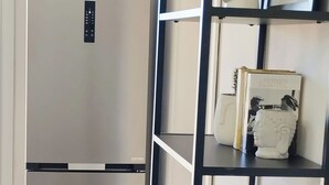 A modern kitchen with a stainless steel refrigerator, a black metal shelving unit, and a white wall with a framed picture and decorative plants.