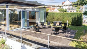 Uma área de estar moderna ao ar livre, com paredes de vidro, piso de madeira e vista para um bairro residencial.