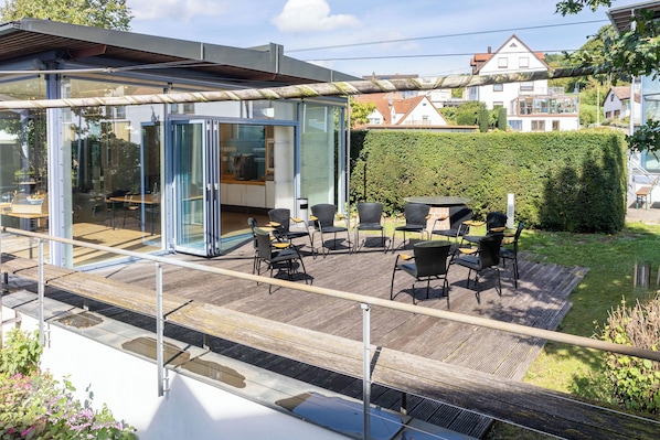 A modern outdoor seating area with glass walls, wooden flooring, and a view of a residential neighborhood.