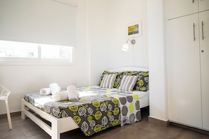 Una cama bien hecha con un edredón estampado, una lámpara de noche y una ventana con persianas.