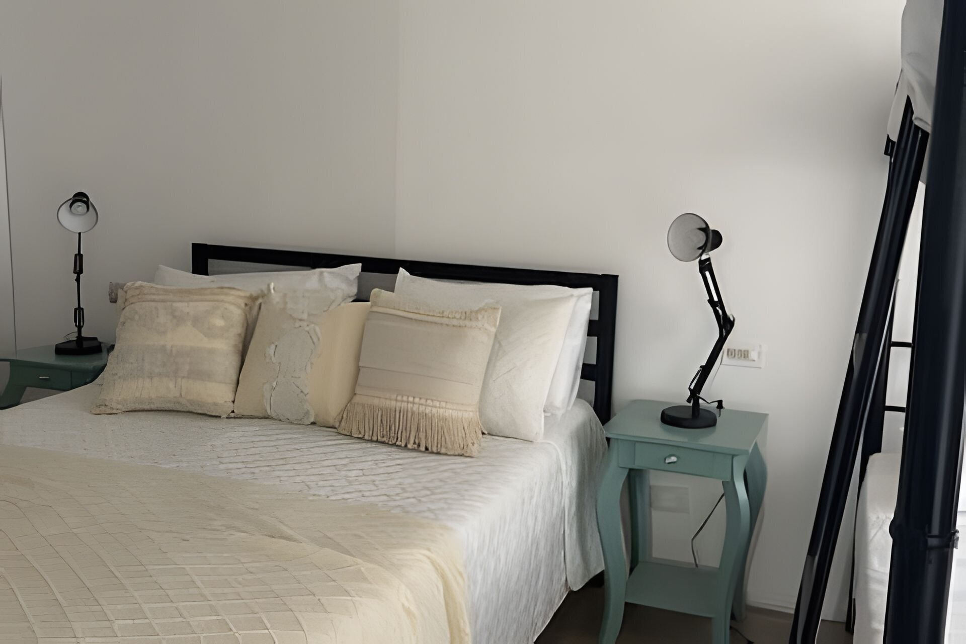 Ein ordentlich bezogenes Bett mit weißer Daunendecke und mehreren Kissen, zwei Nachttische mit Lampen und eine schlichte Wand.