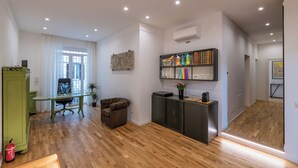 Una sala de estar moderna con piso de madera, un mueble verde, una silla de oficina negra, un sofá marrón y una estantería con objetos de colores.