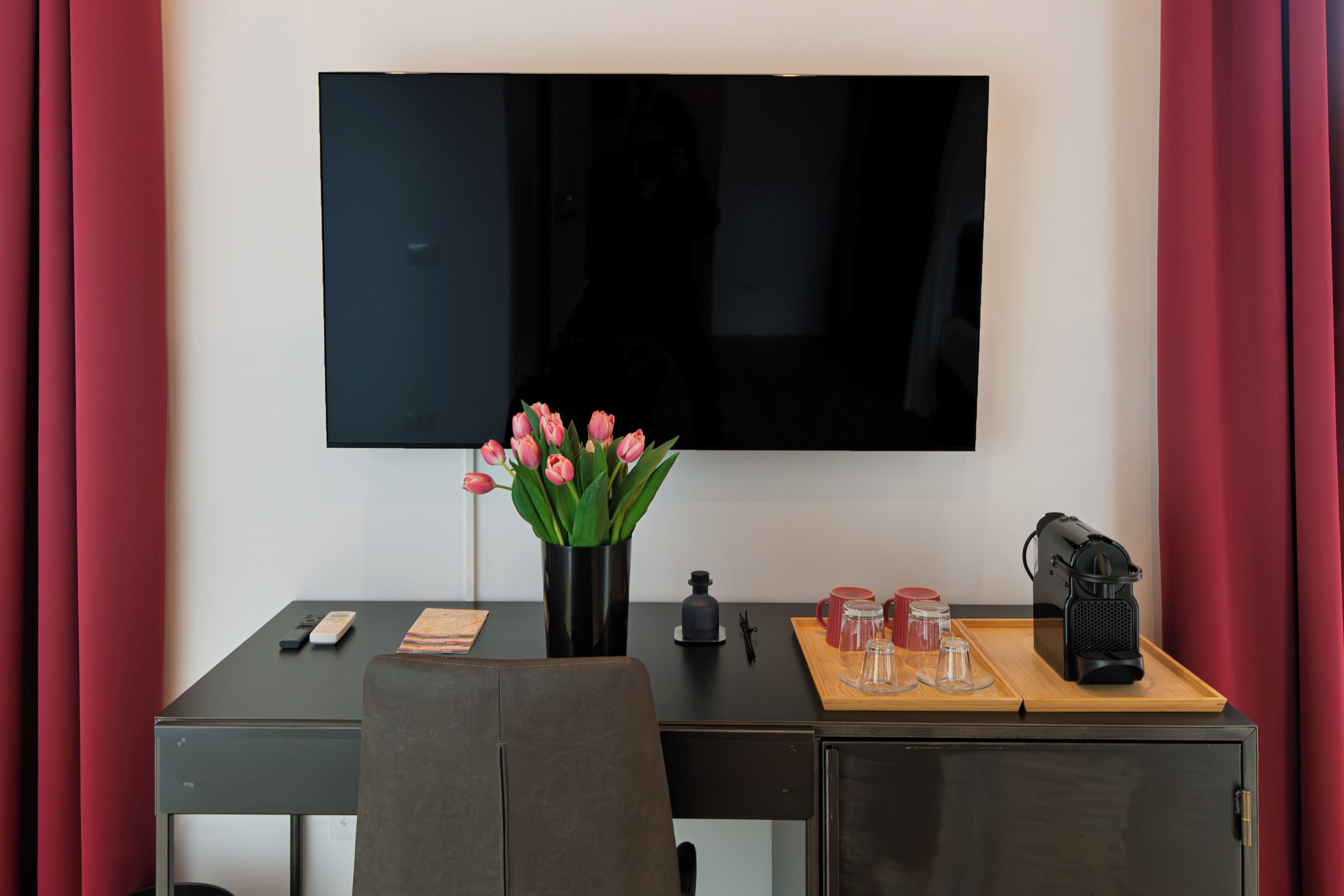 A hotel room with a desk, a flat-screen TV mounted on the wall, a vase with pink flowers, a coffee maker, and a tray with glasses and a cup.