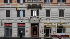 Un edificio con entrada central, dos escaparates visibles con rótulos y un asta con bandera.