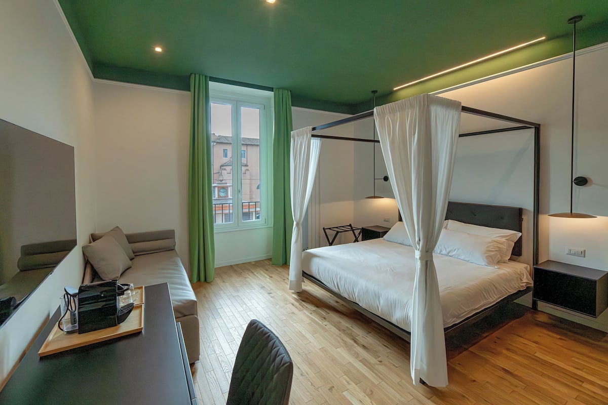 A modern bedroom with a four-poster bed, a sofa, a desk with a tray, and a view of the city through the window.