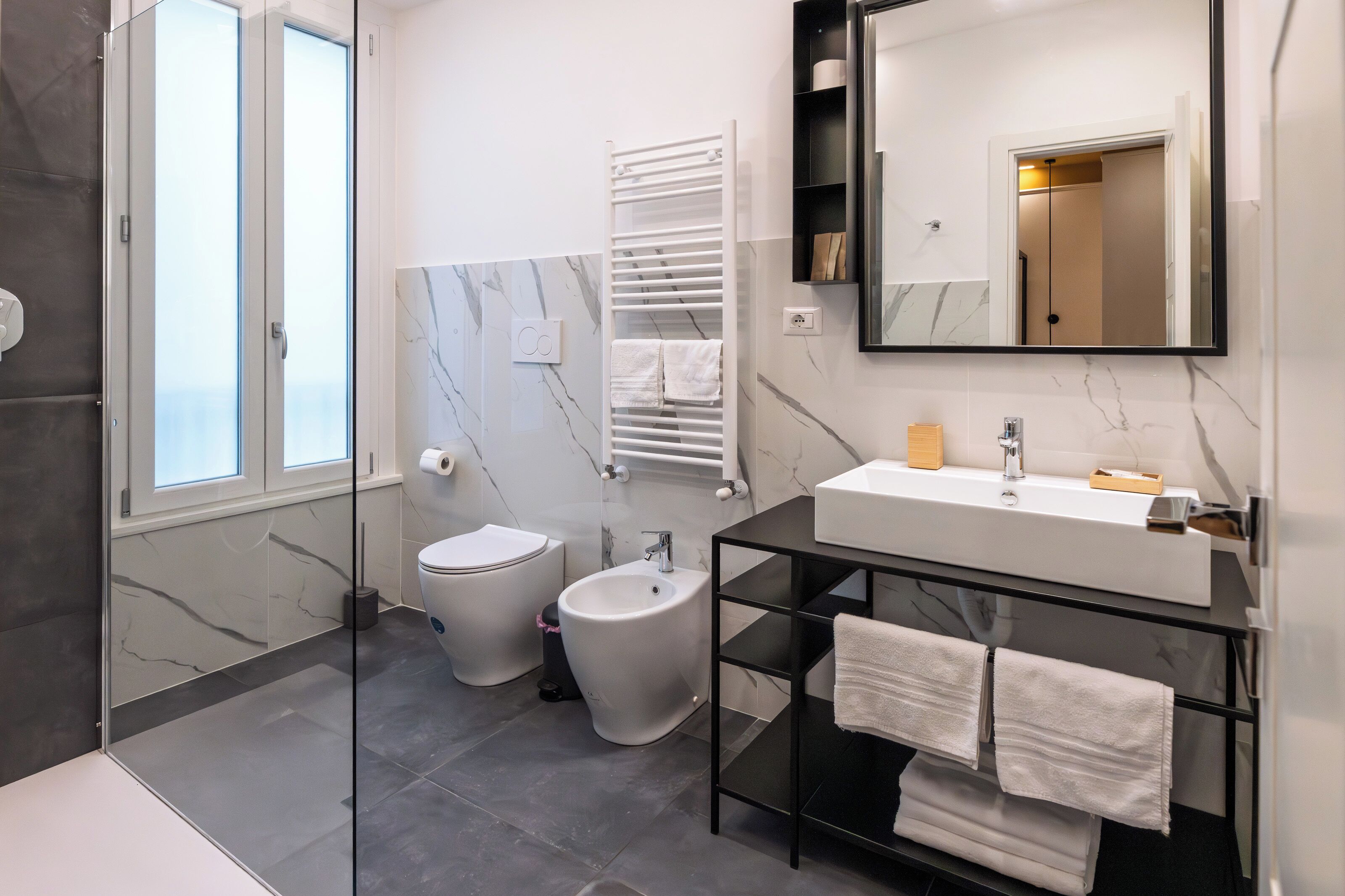 A modern bathroom with a glass shower enclosure, a toilet, bidet, and a vanity with a mirror and towel rack.