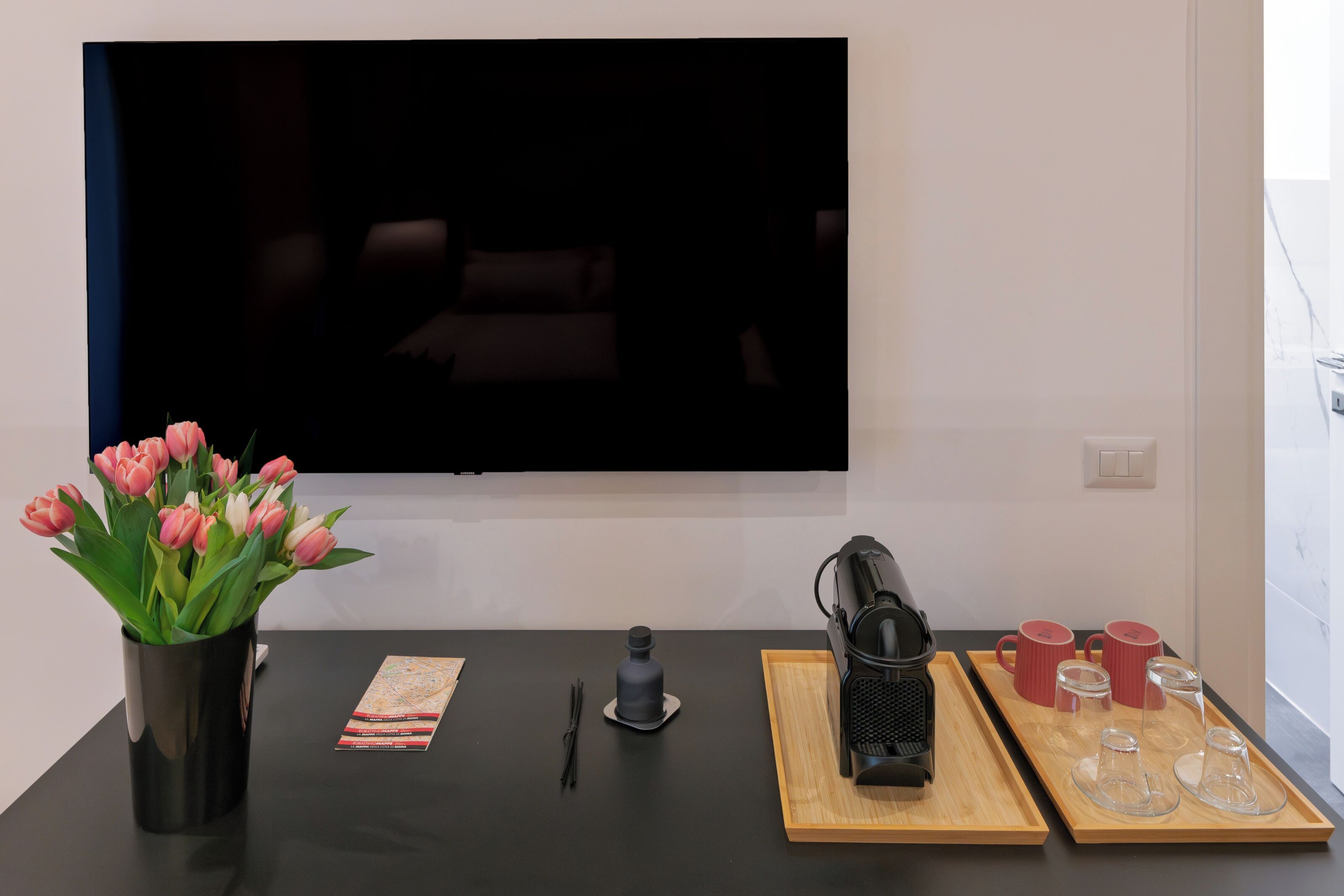 A modern living room with a flat-screen TV mounted on the wall, a coffee table with a tray, a vase of pink tulips, and a coffee machine.